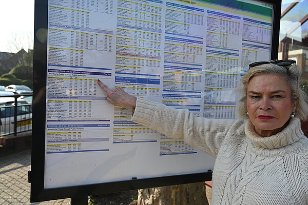 Rachel Gilmour MP standing in front of a Bus Timetable, pointing at the times, whilst looking at the camera